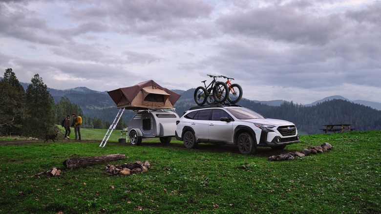 A white Subaru Outback towing a trailer with a tent and a bike, front 3/4 view, couple behind the tent