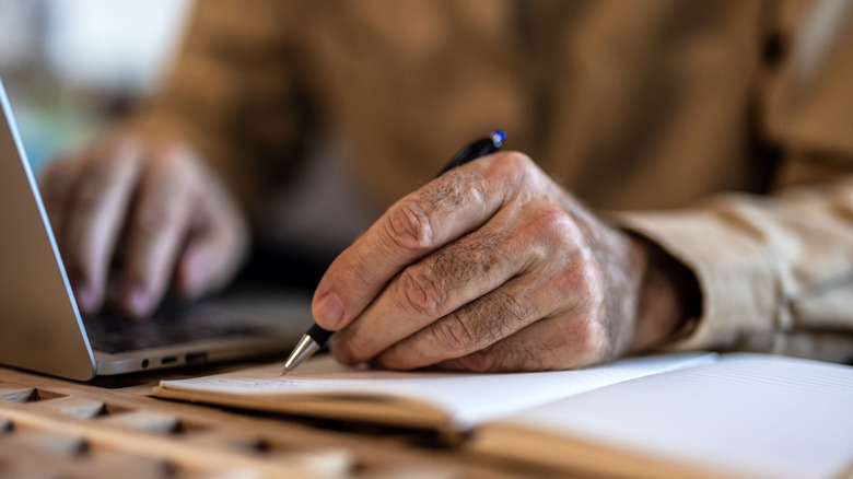Close-up of hands using a laptop and writing in a notebook.