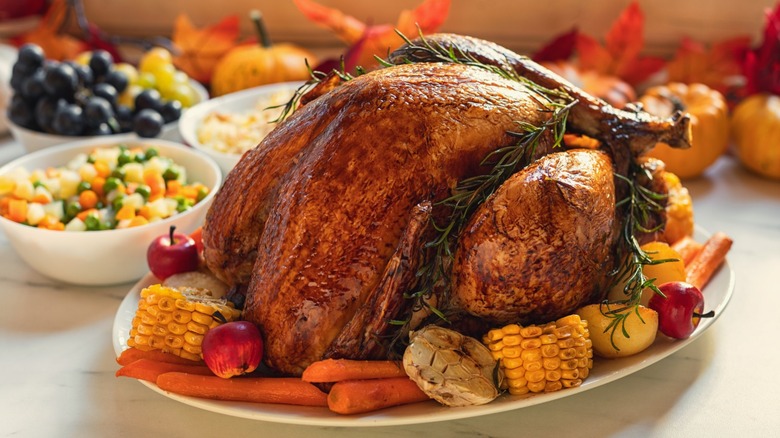 Roasted turkey garnished with rosemary, surrounded by corn, carrots, garlic and small apples on a platter