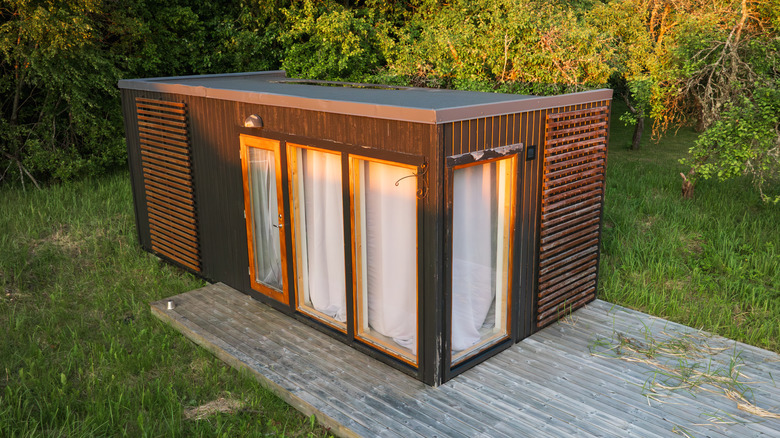 A diagonal view of a tiny home with a deck in a wooded back yard