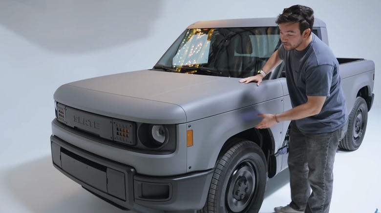 A man gestures toward the gray composite body panel on a Slate Truck displayed in a white studio