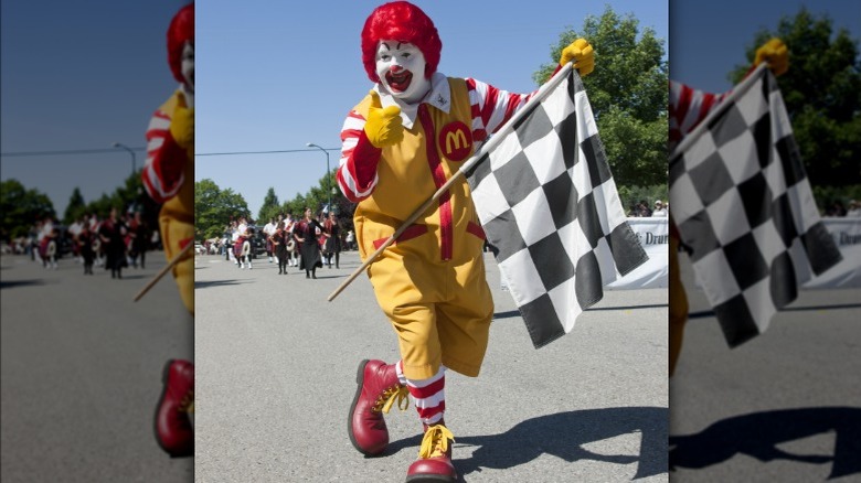 The iconic Ronald McDonald flashes a gloved thumbs up with one hand and holds a racing flag in the other at an organized run/walk event outside.
