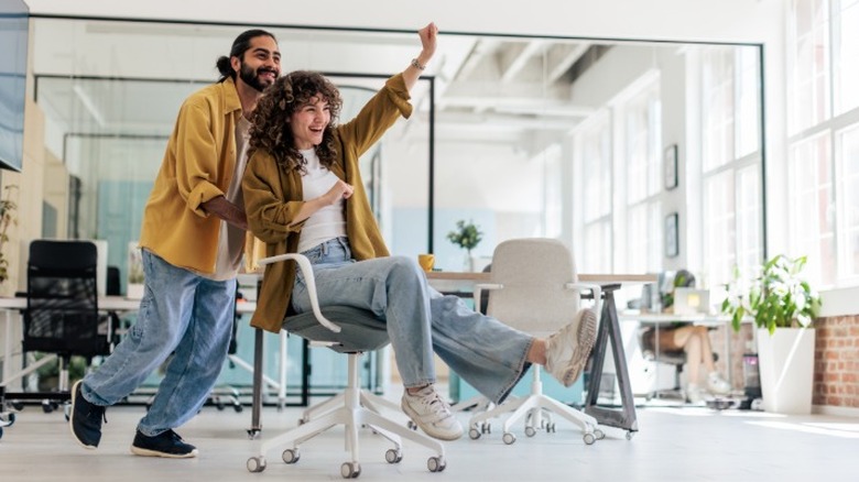Two Gen Z coworkers in an office with one pushing the other in a rolling chair in celebration.