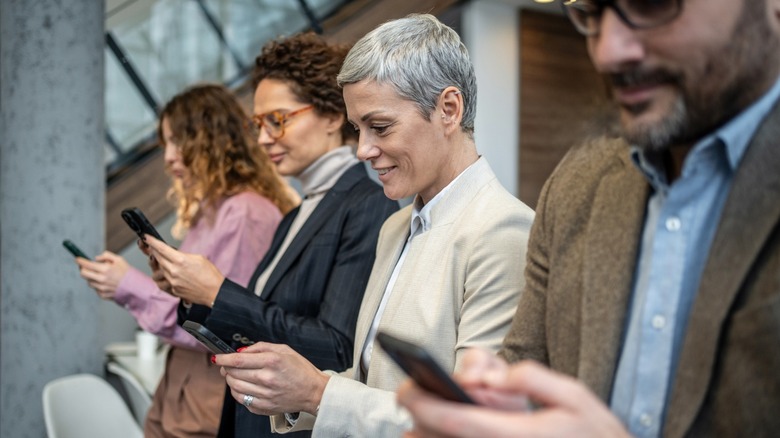 People of different ages standing in a row and looking at finances on their phones.