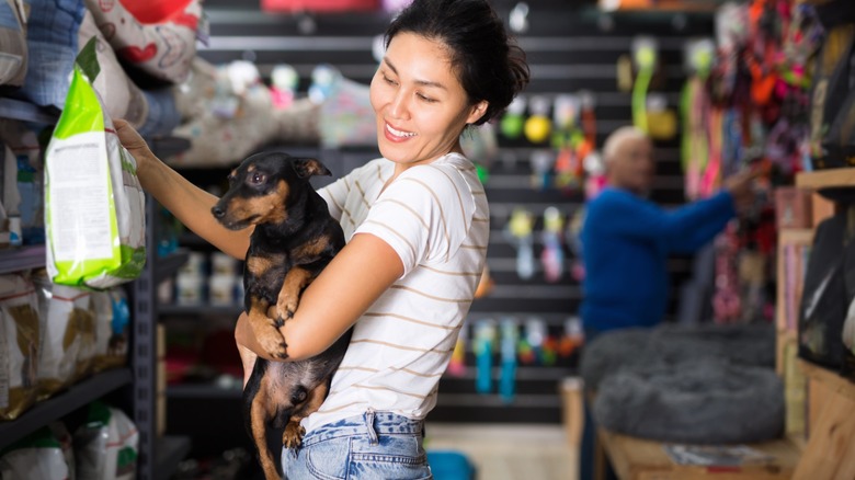 A woman holds a small black-and-tan dog while shopping for pet food in a pet store aisle