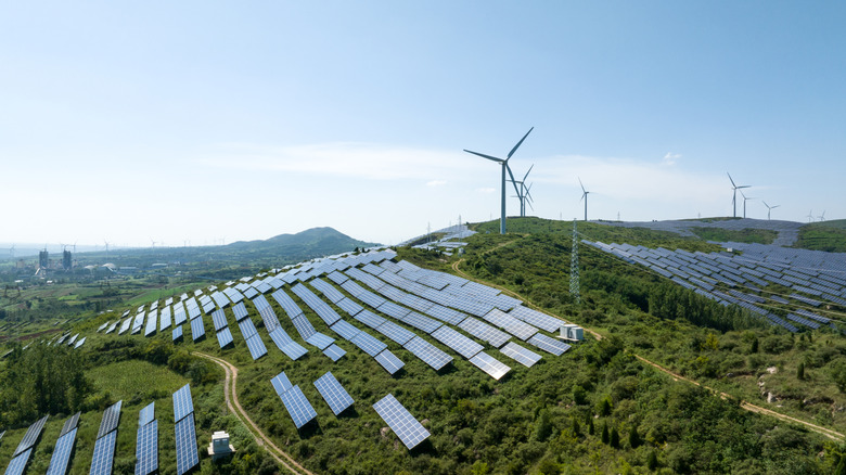 A hilly field filled with solar panels and some wind turbines on a bright day