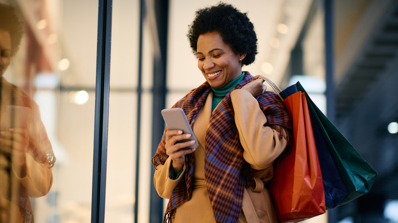 Woman looking at smartphone while holding shopping bags outside a store window