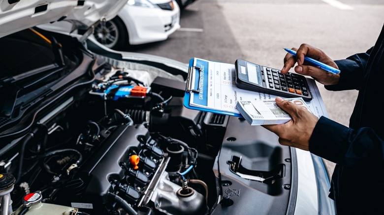 A mechanic uses a calculator while holding a car maintenance checklist beside an open engine bay