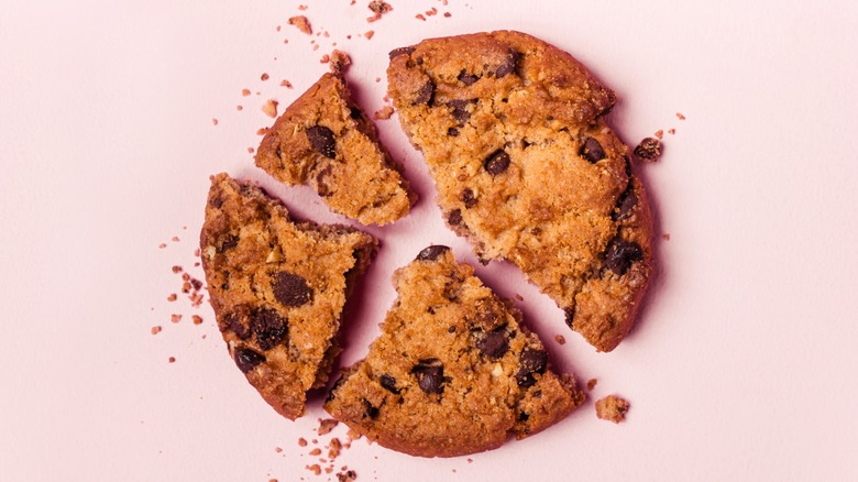 A large chocolate chip cookie on a pink surface smashed into several different pieces.