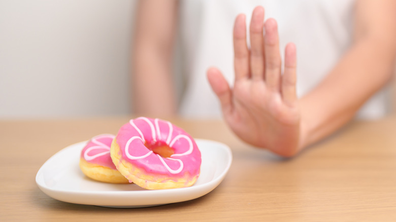 A plate of pink donuts in front of a person in a white shirt, holding up a hand in protesting, rejecting the treats