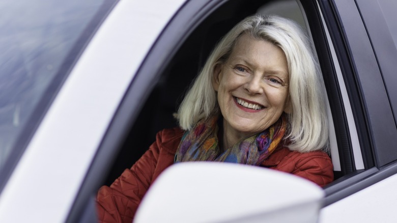 Smiling older woman sitting in the driver's seat of a car, leaning toward the open window while wearing a red jacket and colorful scarf