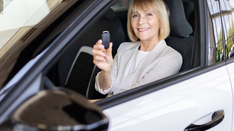 Older woman smiling from the driver's seat of a white SUV, with the window down and the side mirror in the foreground