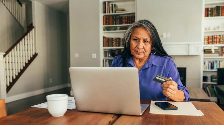 An older woman holding a credit card and looking at her laptop white seated at home.