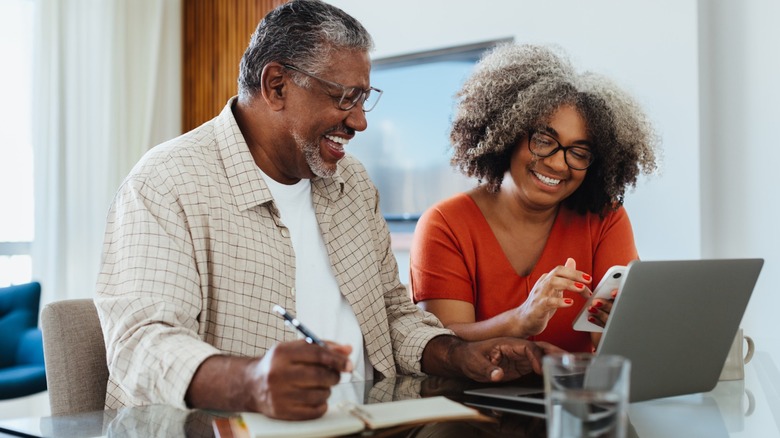 Older couple smiling while reviewing finances on a laptop and smartphone at a table