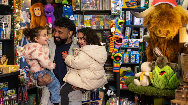 A smiling young man holds his daughters in a toy store