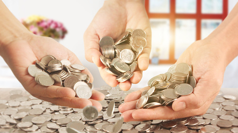 A closeup of three hands holding coins over a large pile of coins on a table.