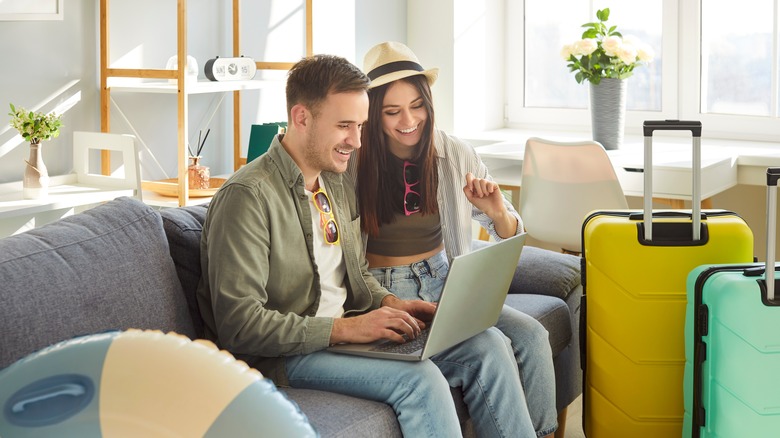 A happy couple sitting on a couch using a laptop with suitcases in front of them.