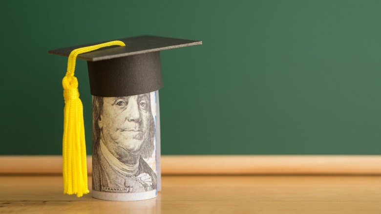 A roll of cash with a graduation cap on top of it sitting on a table in front of a school chalkboard.