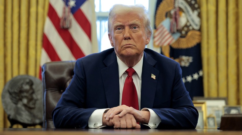 Donald Trump sitting at his desk in the Oval Office with his hands folded.