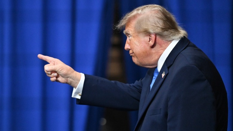 President Donald Trump pointing to the side during a rally in front of blue drapes.
