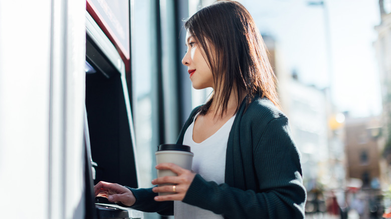 A Millennial woman with a calm demeanore using an ATM while holiding a coffee.