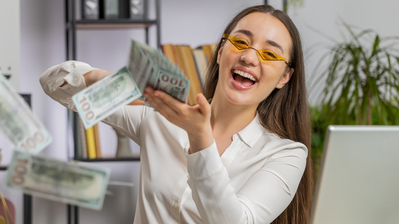 An ecstatic Gen Z woman sitting at a desk throwing cash in the air.