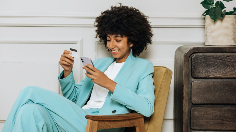 A smiling Gen Z woman looking at her phone and holding a credit card.