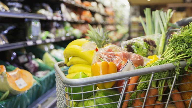 Shopping cart filled with bananas, peppers, tomatoes, carrots, and other produce in a grocery store aisle