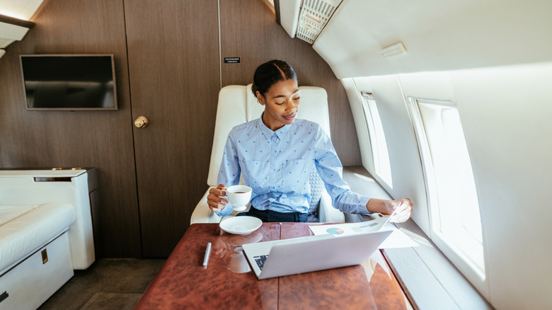 Young woman on a private jet drinking coffee and looking at charts with a laptop open