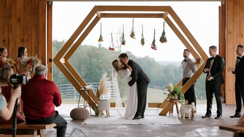 A bride and groom kissing in a farmhouse while their wedding party looks on.