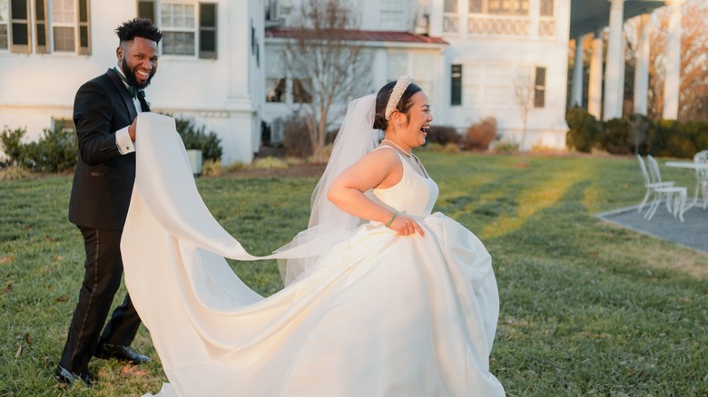 A happy bride walking outside while the groom holds her bridal train.