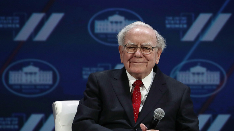 Warren Buffett grinning while holding a microphone on stage during an interview
