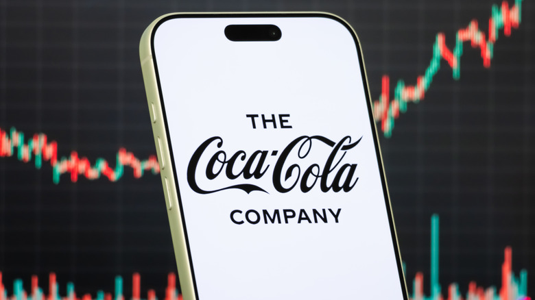 A smartphone with "The Coca-Cola Company" on the screen in black letters against a white background and a stock market graph behind it.