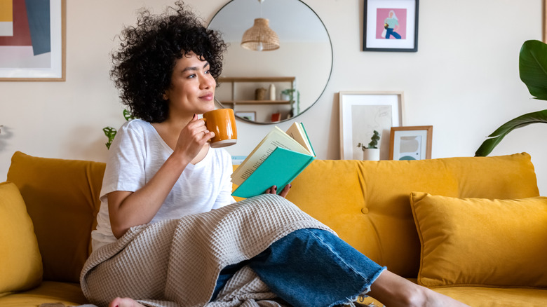 barefoot woman on couch holds a book and mug while looking off into the distance