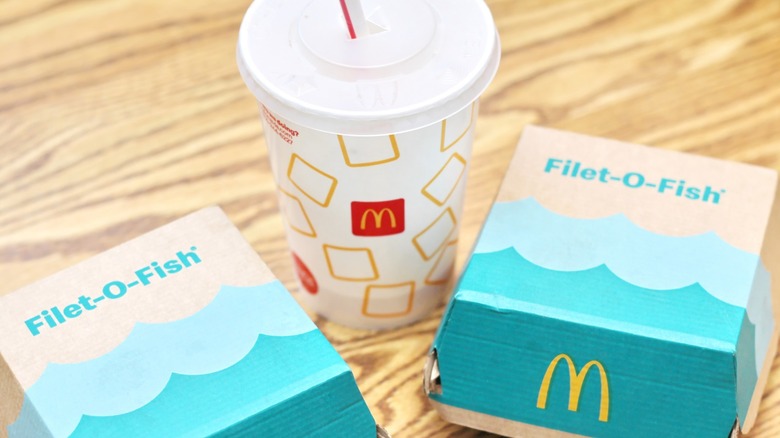 Two McDonald's Filet-O-Fish boxes beside a soft drink on a wooden table.