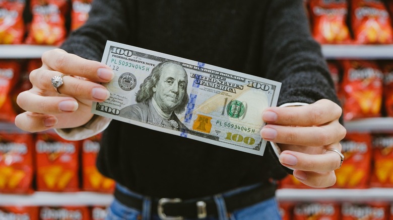 A shopper holds a $100 bill in front of grocery store shelves stocked with packaged goods