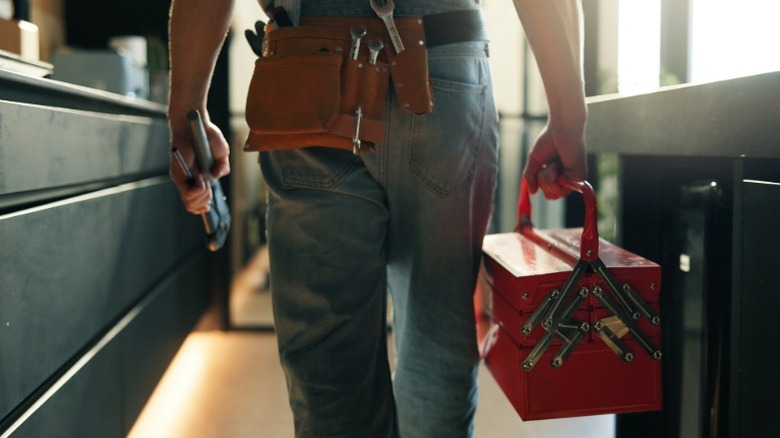 A contractor wearing a tool belt walking in a home with a red toolbox in one hand and pliers in the other.