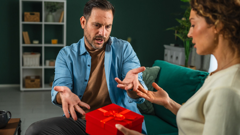 Man and woman seated on couch argue over box with ribbon tied on top