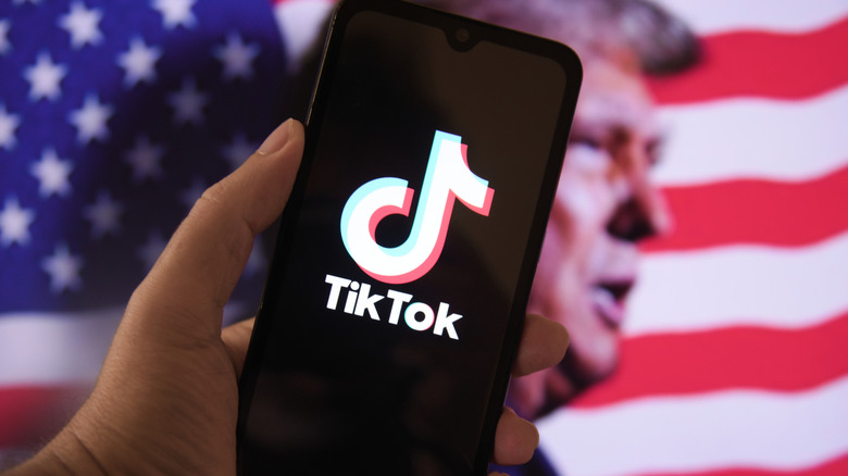 A hand holds up a mobile phone with the TikTok logo in front of a blurred image of Trump and the US flag