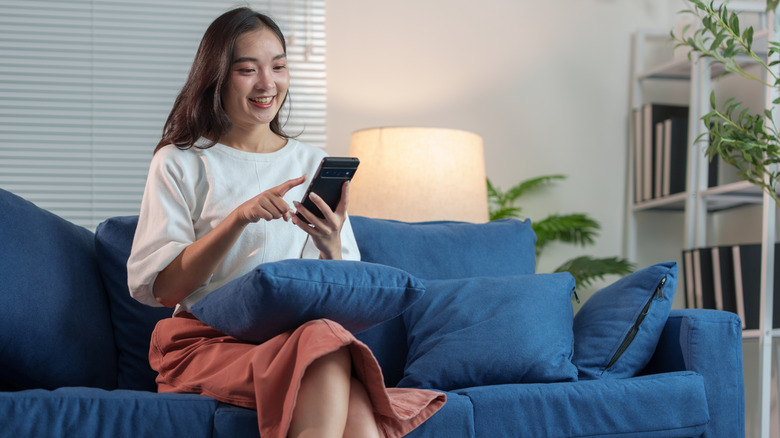 Relaxed woman with pillow in her lap sits on her sofa while using her smartphone