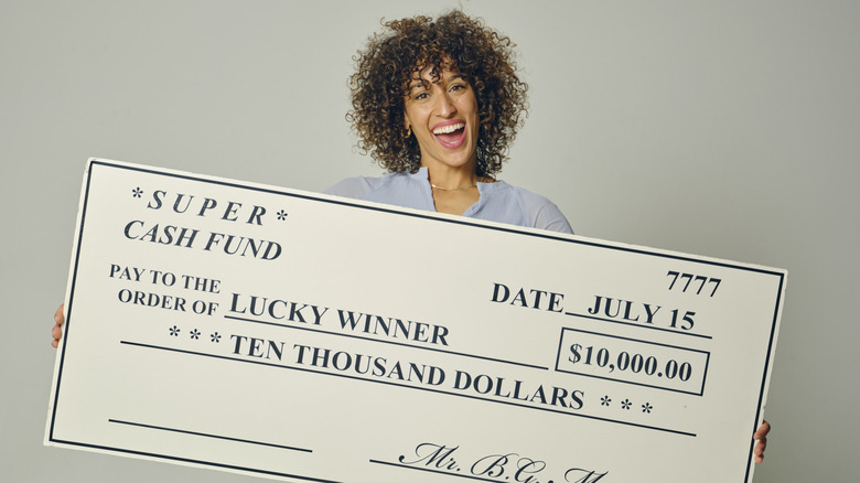 A happy young woman in a studio holding a giant prize check against a gray background.