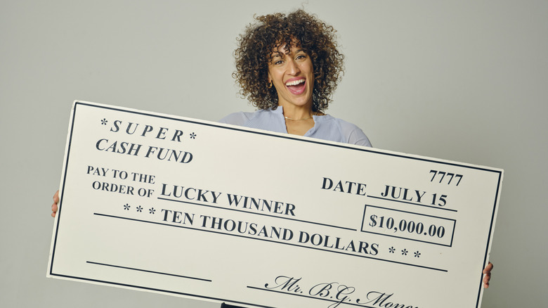 A happy young woman in a studio holding a giant prize check against a gray background.