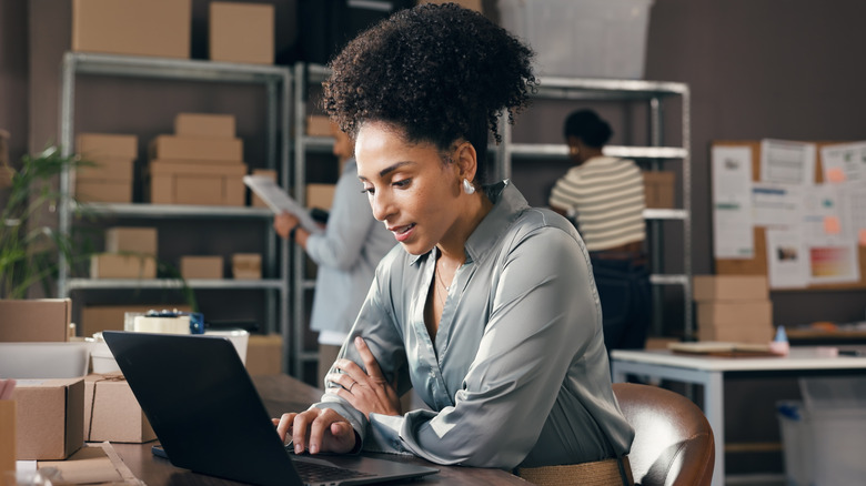 Woman sits in front of laptop while workers load boxes on shelves in background