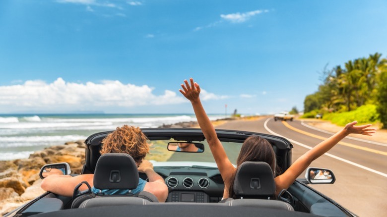 A young man and woman in a convertible driving down a sunny highway with the woman in the passenger seat throwing her arms in the air.