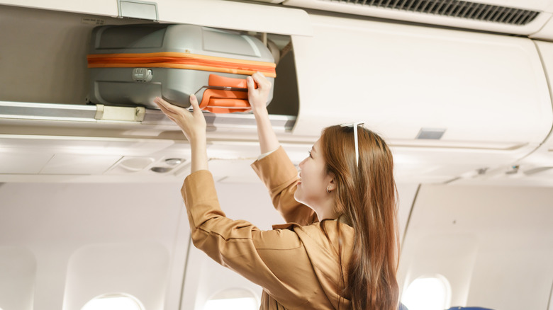 A young woman puts a carry-on suitcase in an airplane's overhead bin