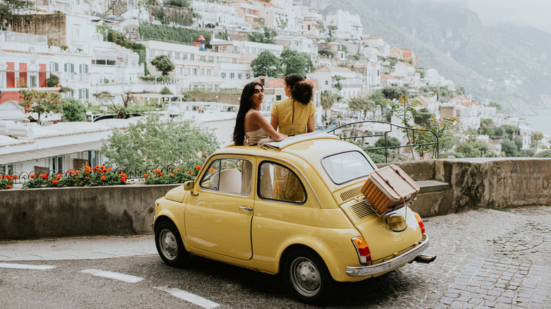 Two women standing out of sun roof in car on European road