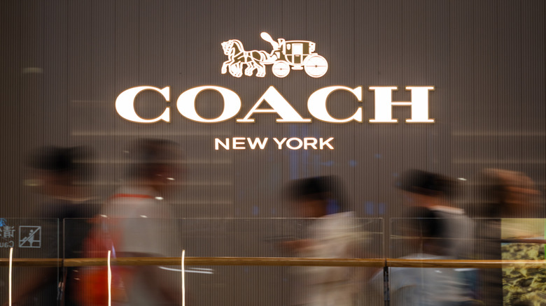 Blurred shoppers hustle underneath an illuminated Coach New York sign in a shopping center