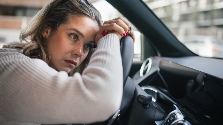 A worried-looking woman rests her head and hands on the steering wheel while sitting in the driver's seat of a car