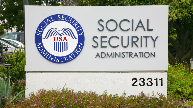 A parking lot sign for a Social Security Administration Field Office, with flowers planted along the bottom and trees in the background