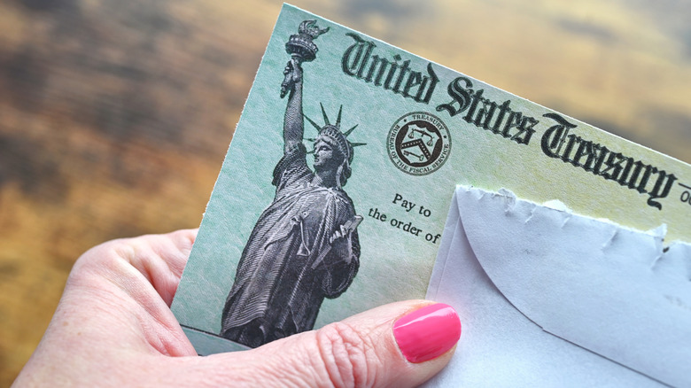 A senior hand with painted pink nails holds a torn envelope and a check from the United States Treasury
